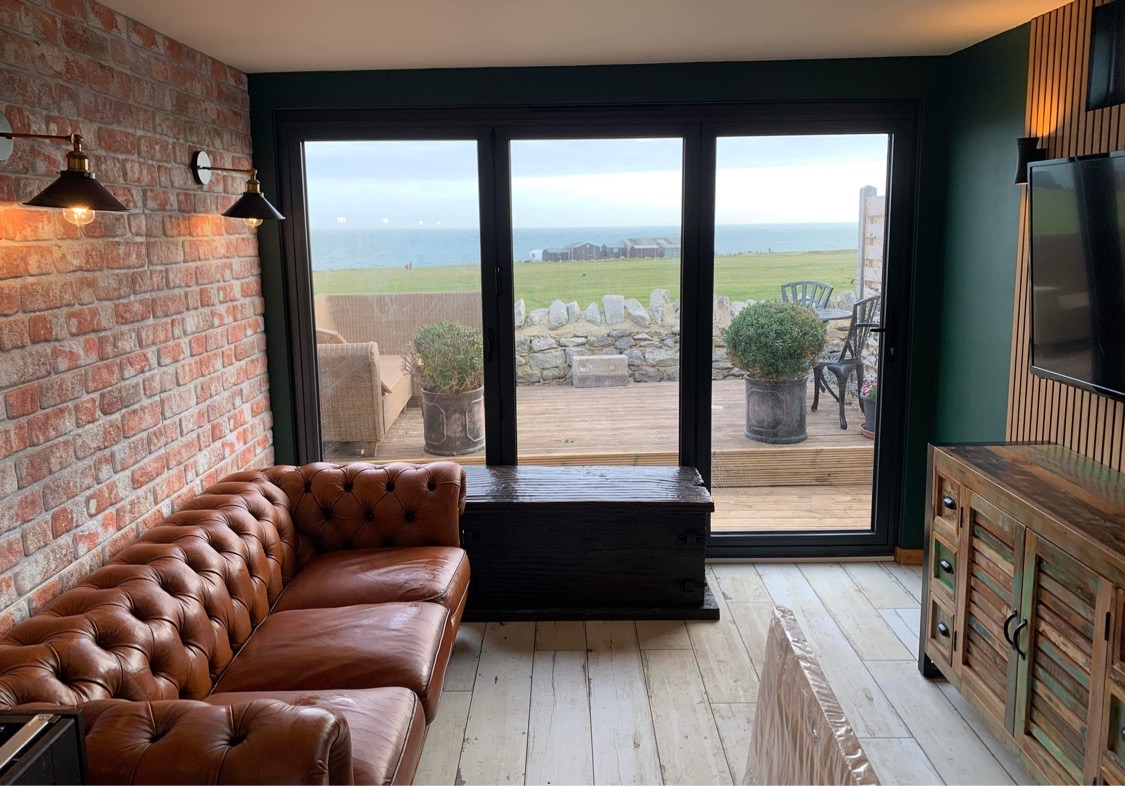 Interior view of a cozy garden room featuring a brown leather sofa, a wooden coffee table, and a decorative wall made of exposed brick, with large sliding doors overlooking a scenic outdoor area.